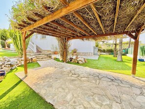 En uteplats med stenläggning och en pergola ovanför, omgiven av en välskött gräsmatta och en stenmur.