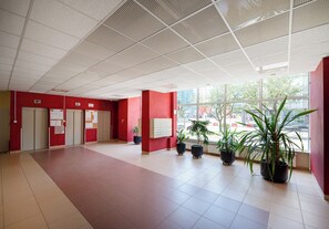 Um hall espaçoso com teto alto, paredes vermelhas e piso ladrilhado. Várias plantas em vasos e uma grande janela com vista para o exterior.