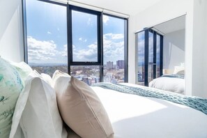 A hotel room with a large window, a bed with white bedding, and a view of the cityscape. - L7 Stunning 2 Bedroom Apt Skyline Views (Broadbeach)