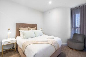 Ein ordentlich eingerichtetes Schlafzimmer mit Bett, Nachttischen, einem Sessel und einem Fenster mit Vorhängen.