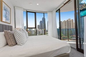 Ein modernes Schlafzimmer mit einem großen Bett, Blick auf die Stadt durch raumhohe Fenster und einem gerahmten Kunstwerk an der Wand.