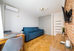 Un espace de vie compact comprenant un canapé, une table à manger avec des chaises, un téléviseur fixé à un mur en briques et un ventilateur de plafond.