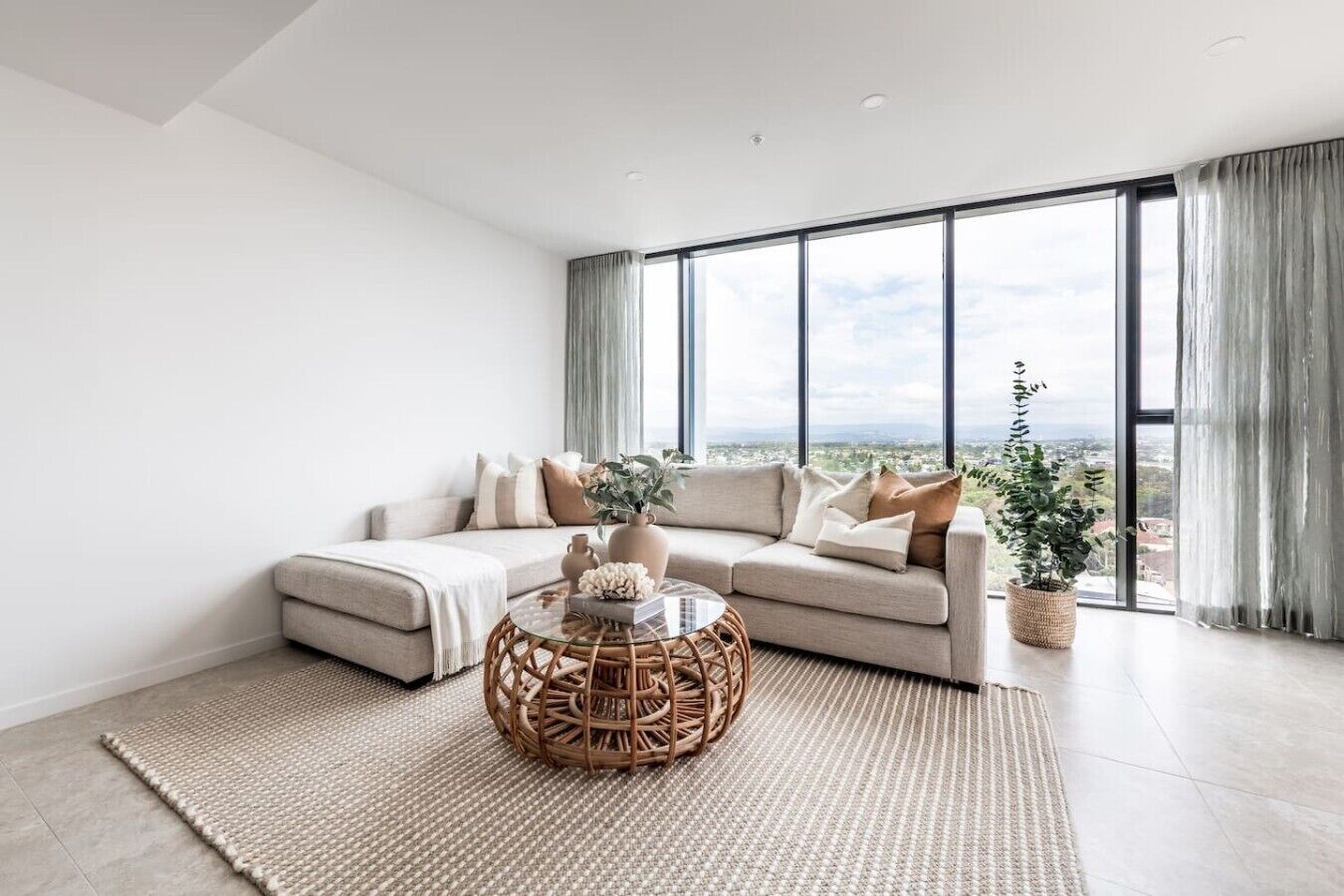 A modern living room with a sectional sofa, a round wicker coffee table, and a large window with a view.