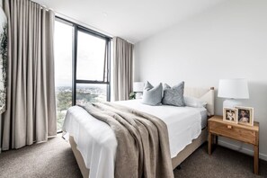 A hotel room with a large bed, a nightstand with a lamp, and a view of a cityscape through the window. - Lv 12 Luxury 3 Bedroom APT Hinterland Views (Broadbeach)