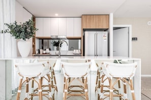 A modern kitchen with white cabinetry, a stainless steel refrigerator, and a central island with white bar stools. - Lv 12 Luxury 3 Bedroom APT Hinterland Views (Broadbeach)