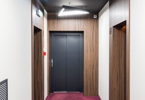 A hallway with a dark elevator door, wooden paneling on the walls, and a red emergency exit sign. - Pawia Studio Centrum 75 (Warszawa)
