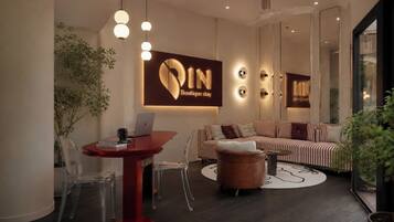 A modern lounge area with a sofa, a round wooden ottoman, a red table with a laptop, and a wall-mounted sign.