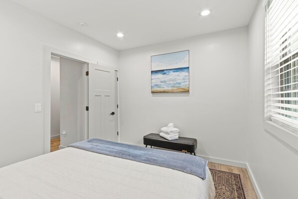 A bedroom with a bed, a bedside table with towels, a painting on the wall, and a window with blinds. - 4BR/3BA Modern GVL Retreat, Fire Pit + Fence (Greenville)