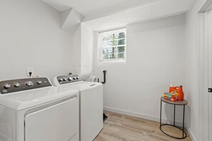Un lavadero con dos lavadoras, una ventana con persianas y una pequeña mesa con productos de limpieza.