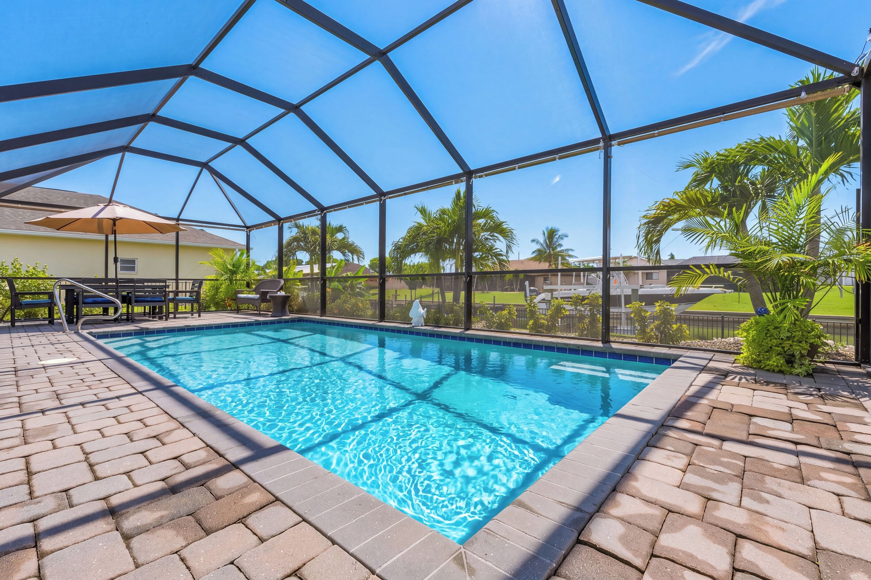 A covered outdoor swimming pool with a clear blue water surface, surrounded by a brick patio, and a view of a residential area with palm trees and houses.