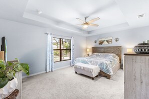 Une chambre à coucher comprenant un lit, un ventilateur de plafond, une fenêtre avec des rideaux, une lampe de chevet et une œuvre d’art accrochée au mur.
