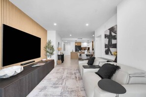 Ein modernes Wohnzimmer mit einem großen Flachbildfernseher, einem dunklen Holz-Konsolentisch, einem weißen Sofa und einer Küche im Hintergrund.