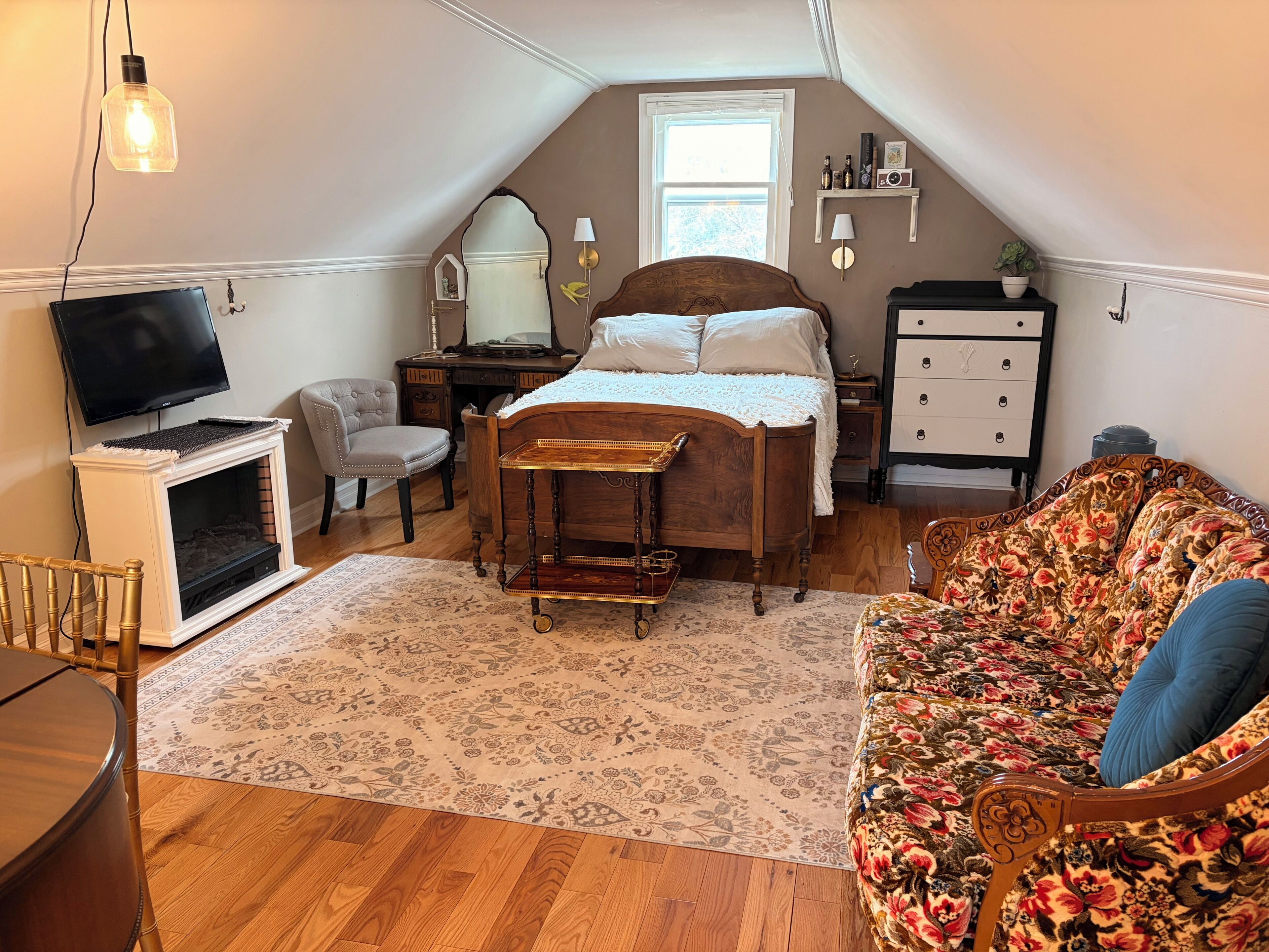 A bedroom with a bed, a dresser, a television, a chair, a sofa, and a fireplace.