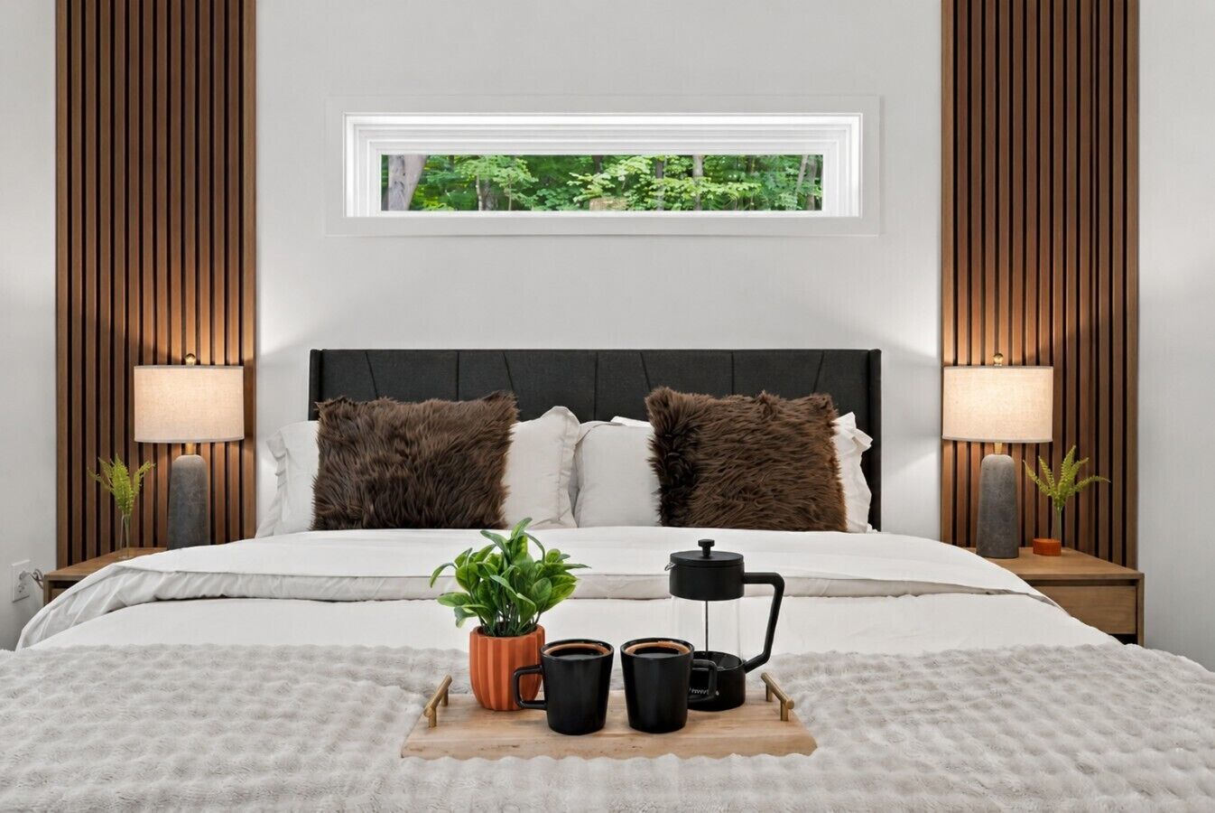 Un dormitorio con cama, dos almohadas, una mesita de noche con lámpara, una ventana con vistas a la vegetación y una bandeja con una planta y tazas.