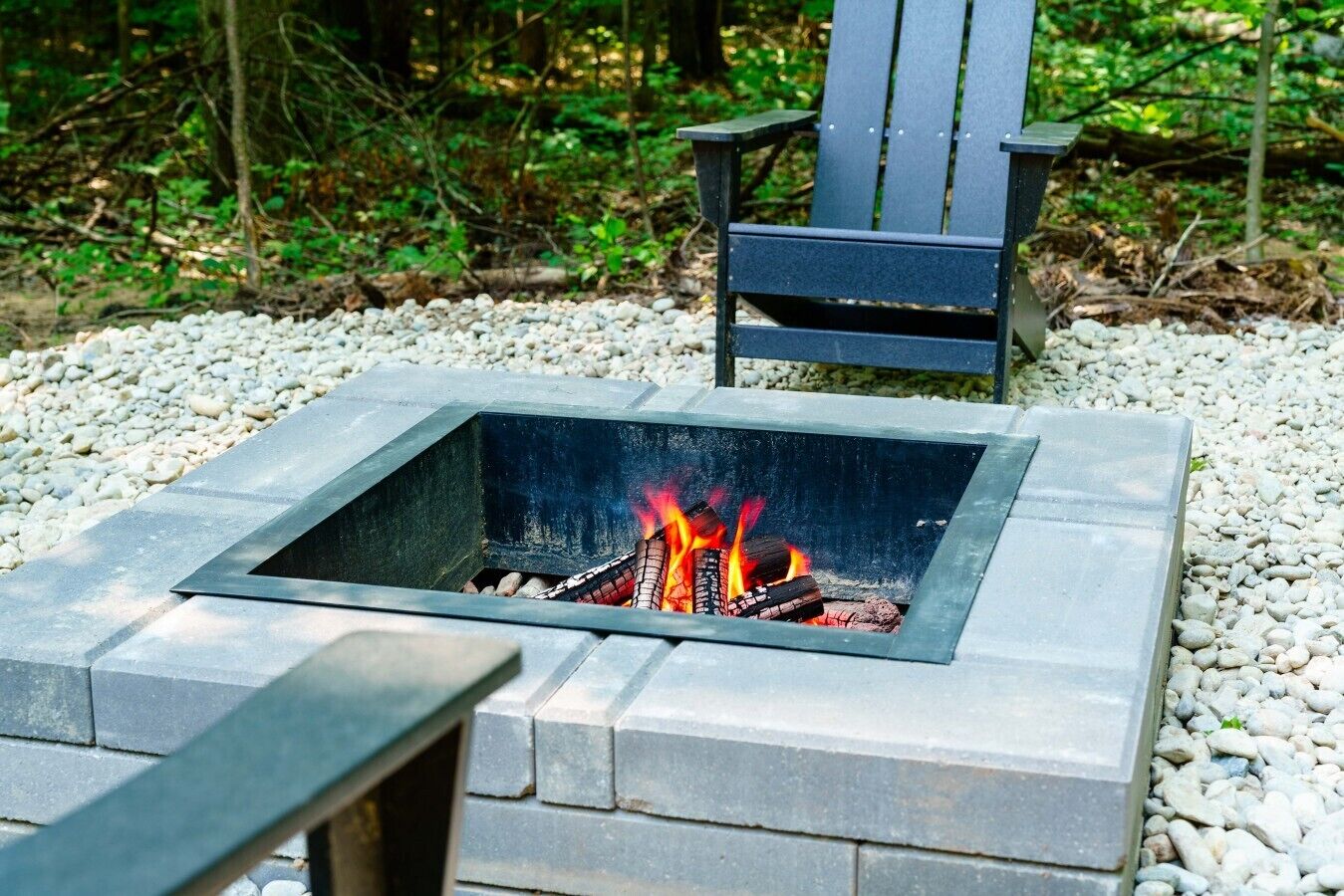 A square fire pit with a metal frame, surrounded by white pebbles, and a wooden chair in the background.