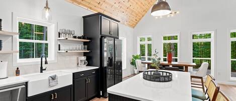 A modern kitchen with black cabinets, a white countertop, and a large island. There are multiple windows providing natural light and views of greenery outside.