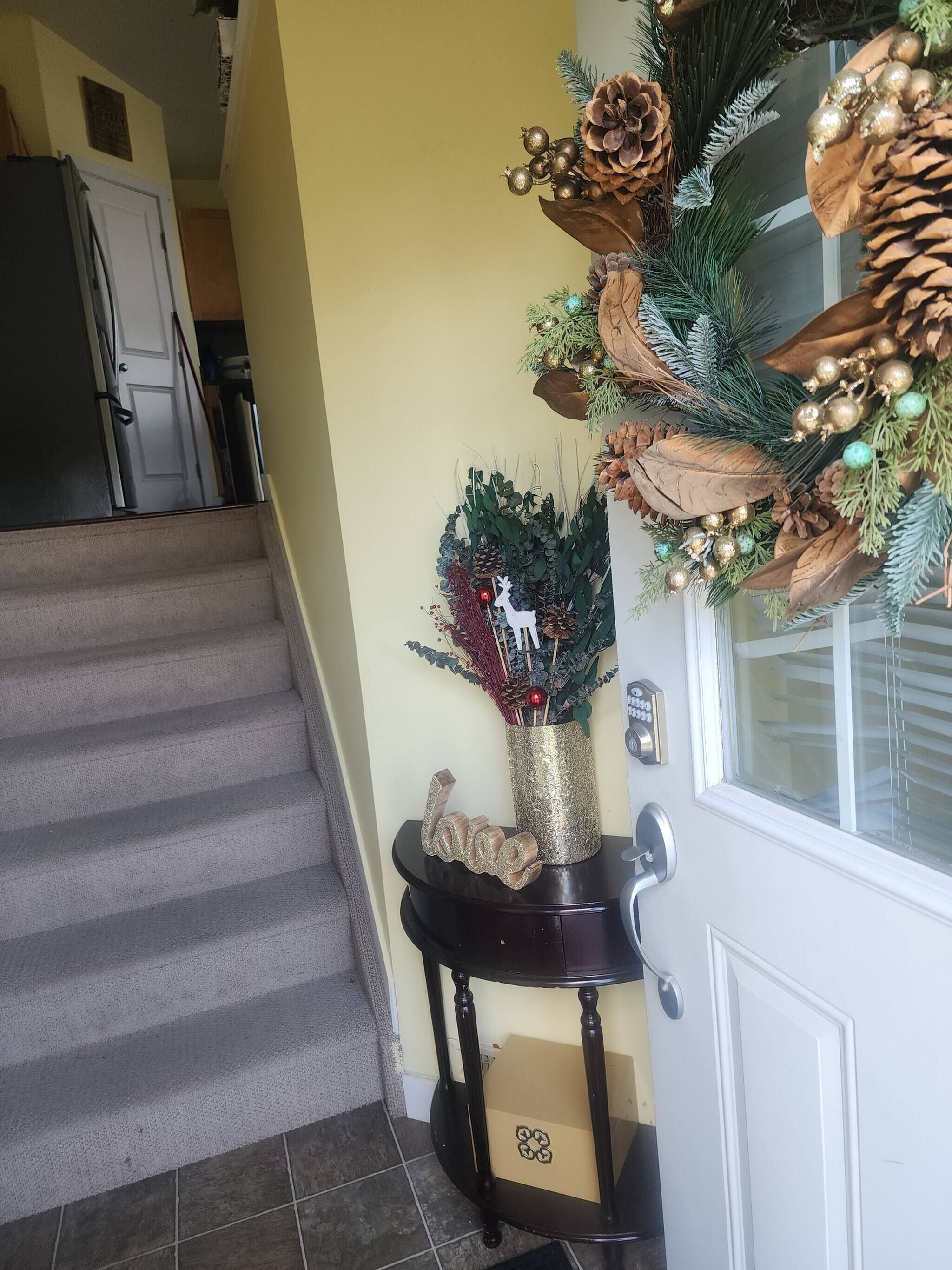 Un escalier menant à une porte ornée d’une guirlande décorative, ainsi qu’une petite table sur laquelle se trouvent un vase et une décoration représentant un renne.