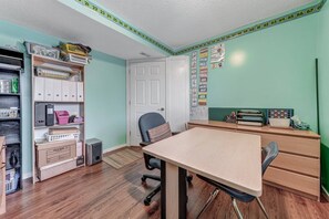 Una habitación con una mesa de madera, sillas, una estantería, un archivador y un tablón de anuncios con material educativo.