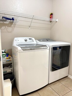 A laundry room with two white front-loading washing machines, a shelf above with a fire extinguisher, and a cart with cleaning supplies. - Stylish 4-bedroom retreat with 3 king beds and twin trundle in charming Frisco. (Frisco)