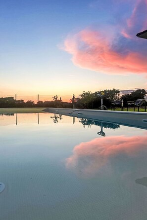Klarer Himmel mit Sonnenuntergangsspiegelung auf einer ruhigen Wasseroberfläche, umgeben von einem Zaun und Bäumen.