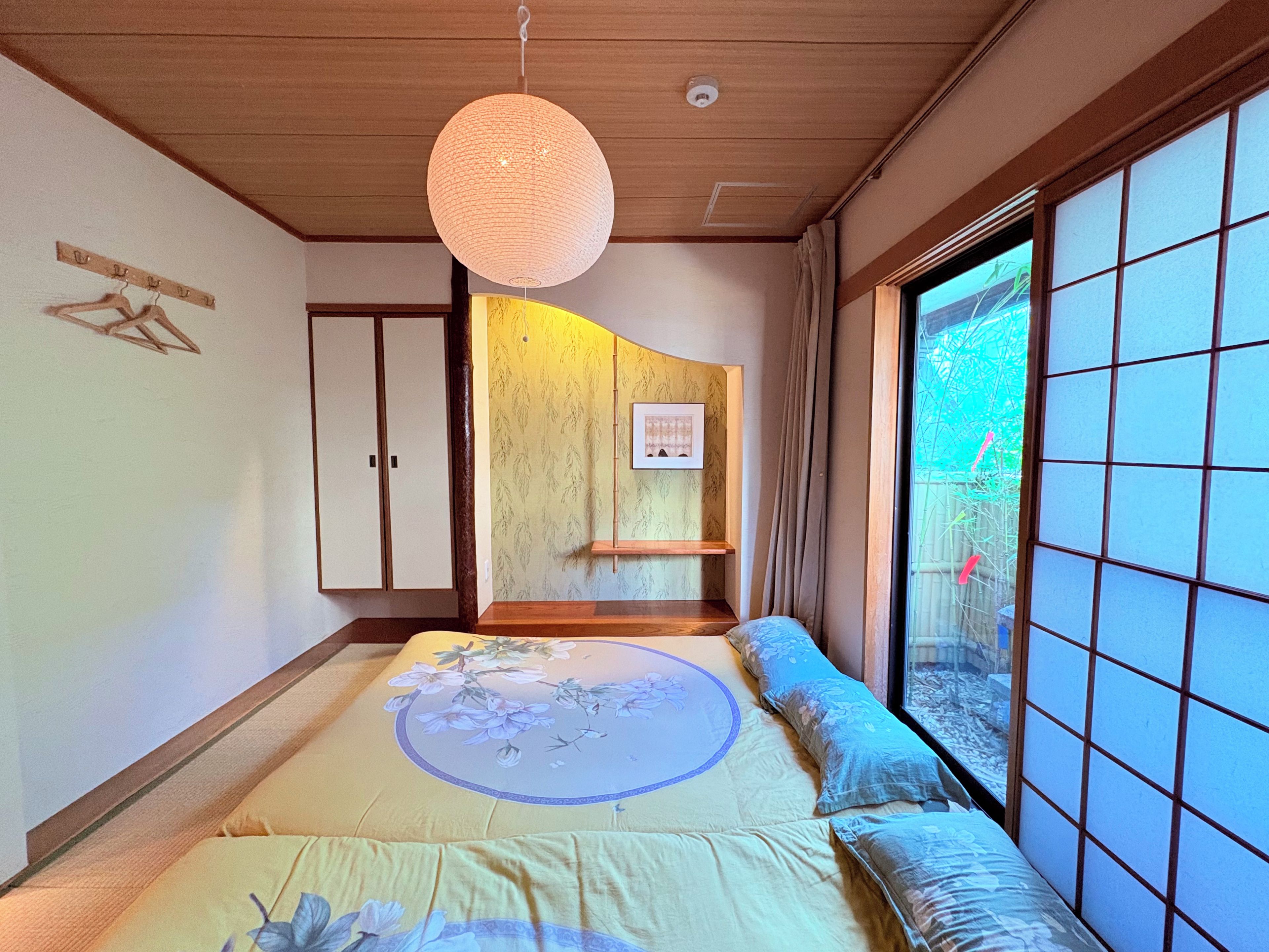 A traditional Japanese-style room with tatami flooring, a low wooden bed with a floral bedspread, a hanging paper lantern, and a sliding shoji screen door.
