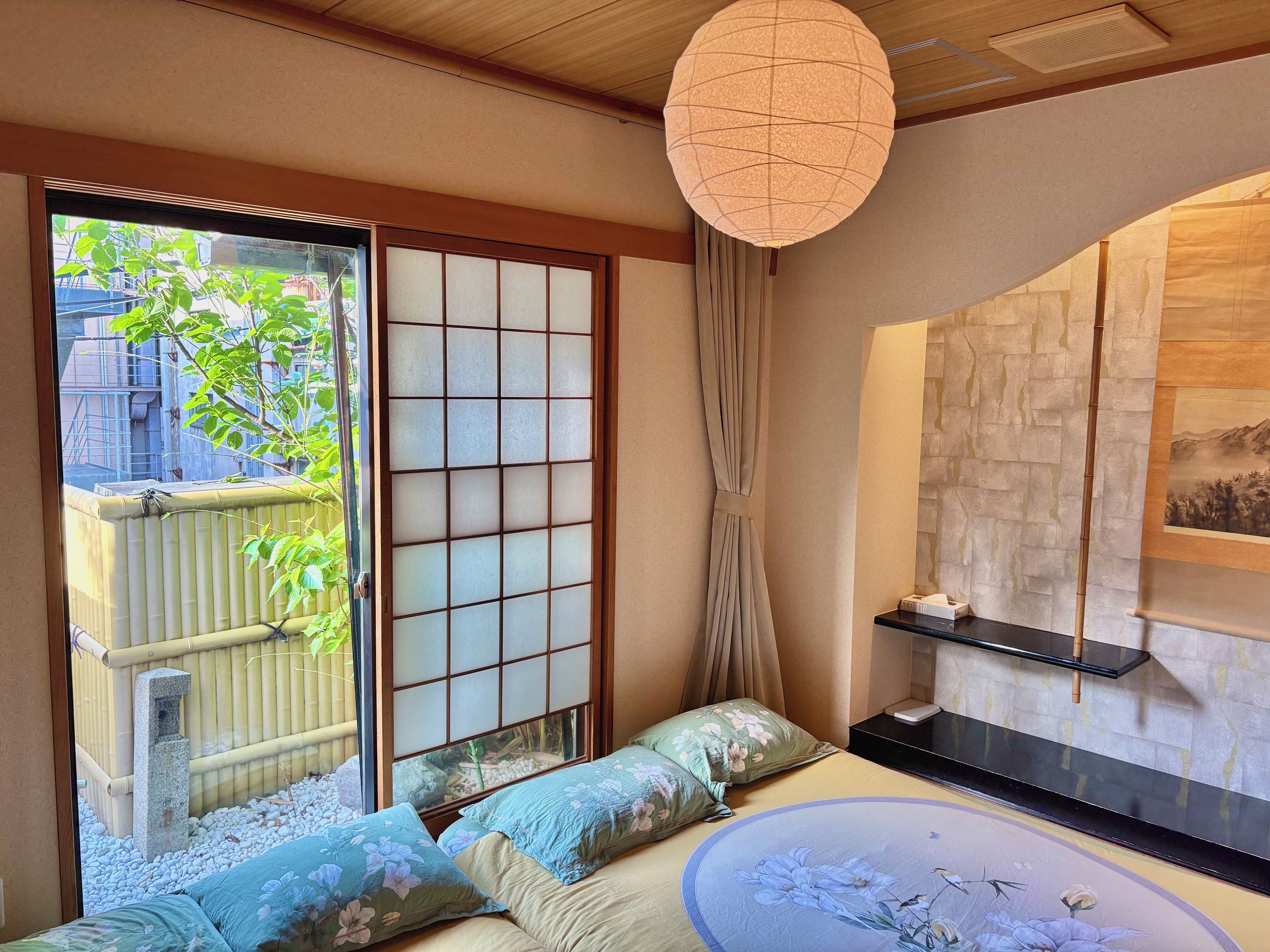 Een traditionele Japanse kamer met tatami vloeren, een shoji scherm, een tatami zitbank met kussens, een hangende papieren lantaarn en uitzicht op groen buiten.