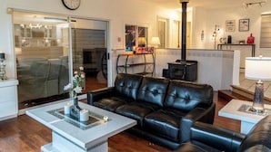 A living room with a black leather sofa, a coffee table, a clock on the wall, and a kitchen area in the background.