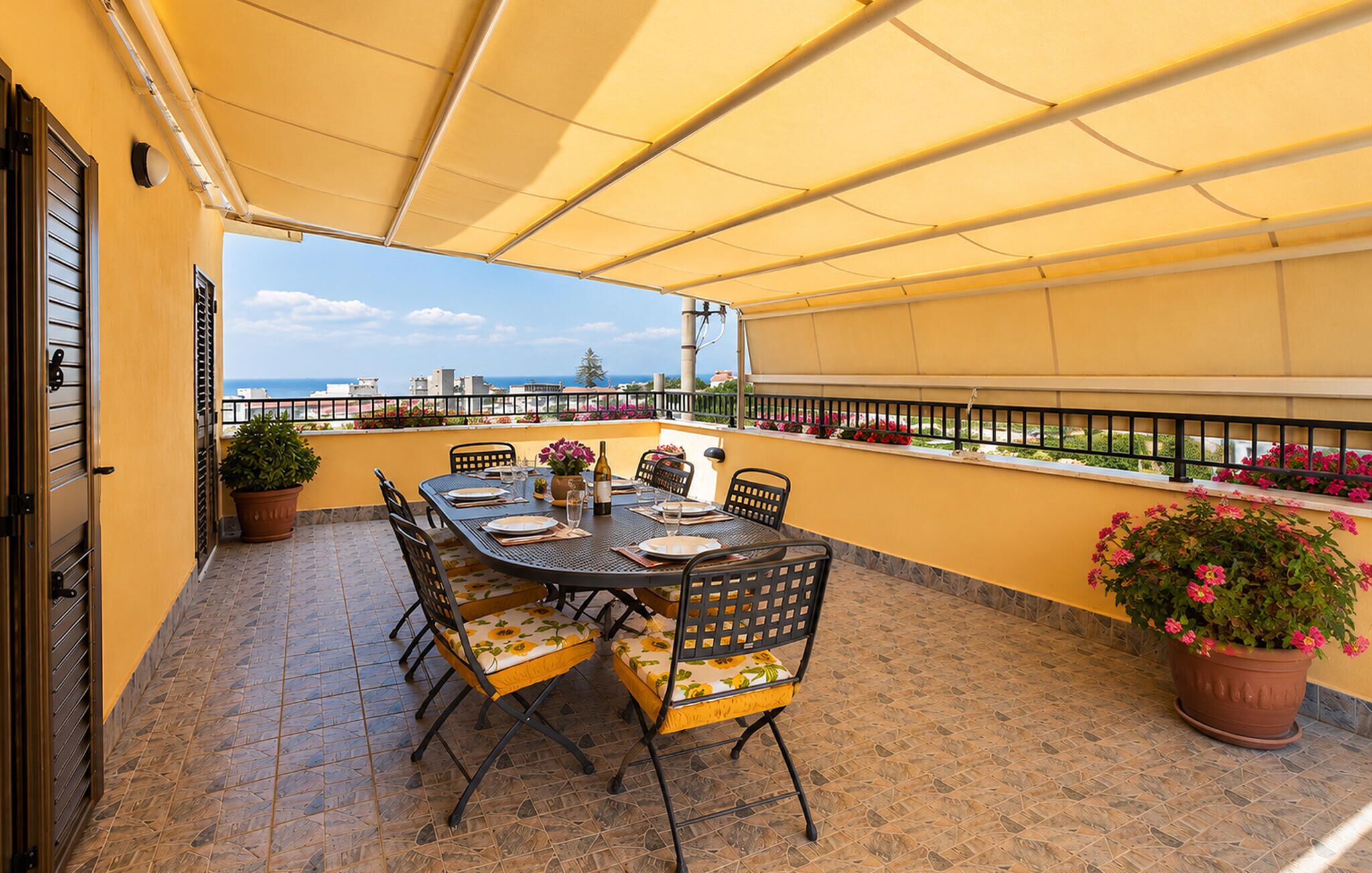 A terrace with a table set for dining, potted plants, and a view of the sea.
