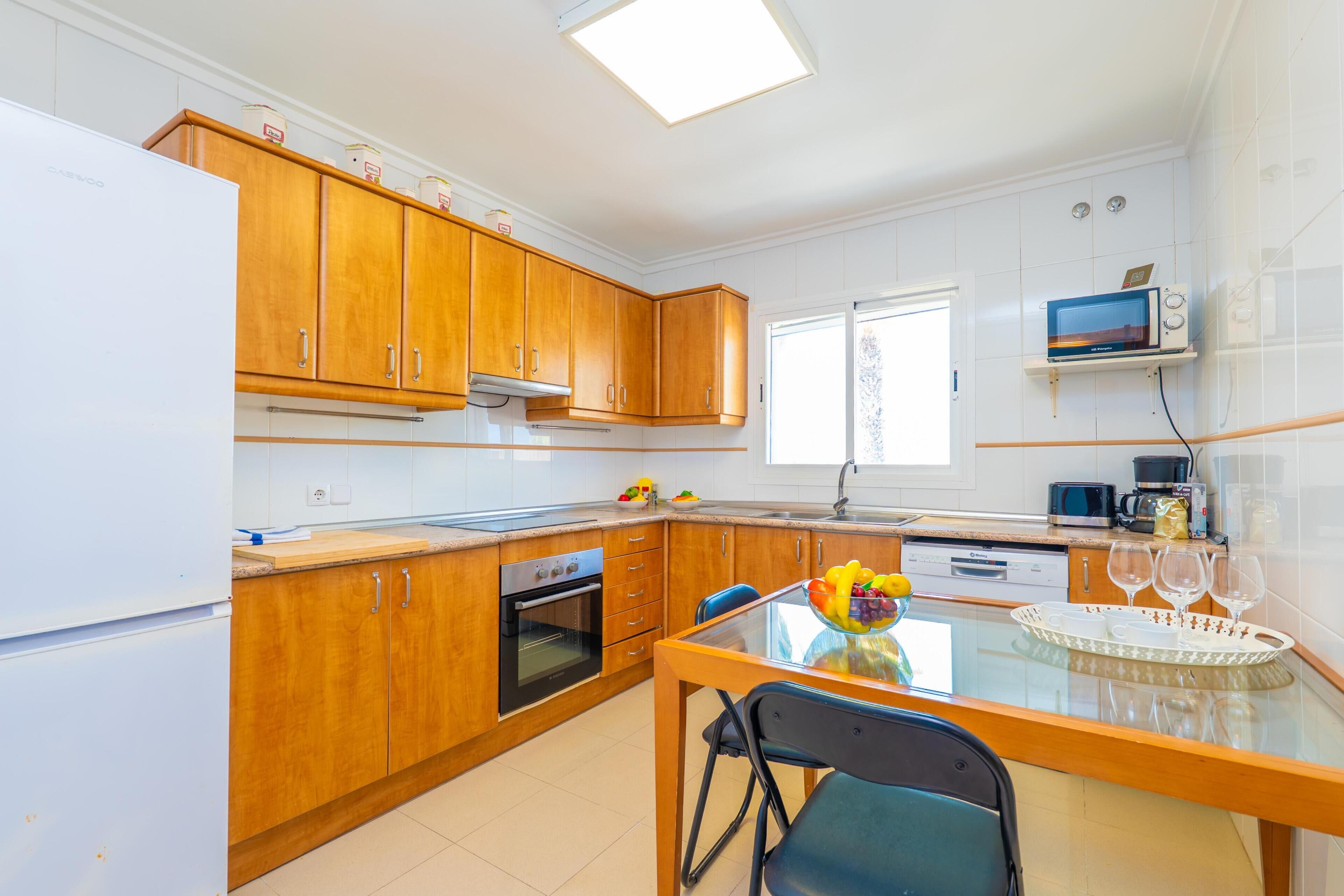 Una cocina moderna con armarios de madera, horno empotrado, microondas y comedor con mesa de vidrio.