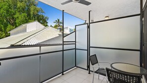 Un balcon doté d’une paroi en verre, d’un ventilateur de plafond, d’une table avec une chaise, et offrant une vue sur un bâtiment dont le toit est en pente.