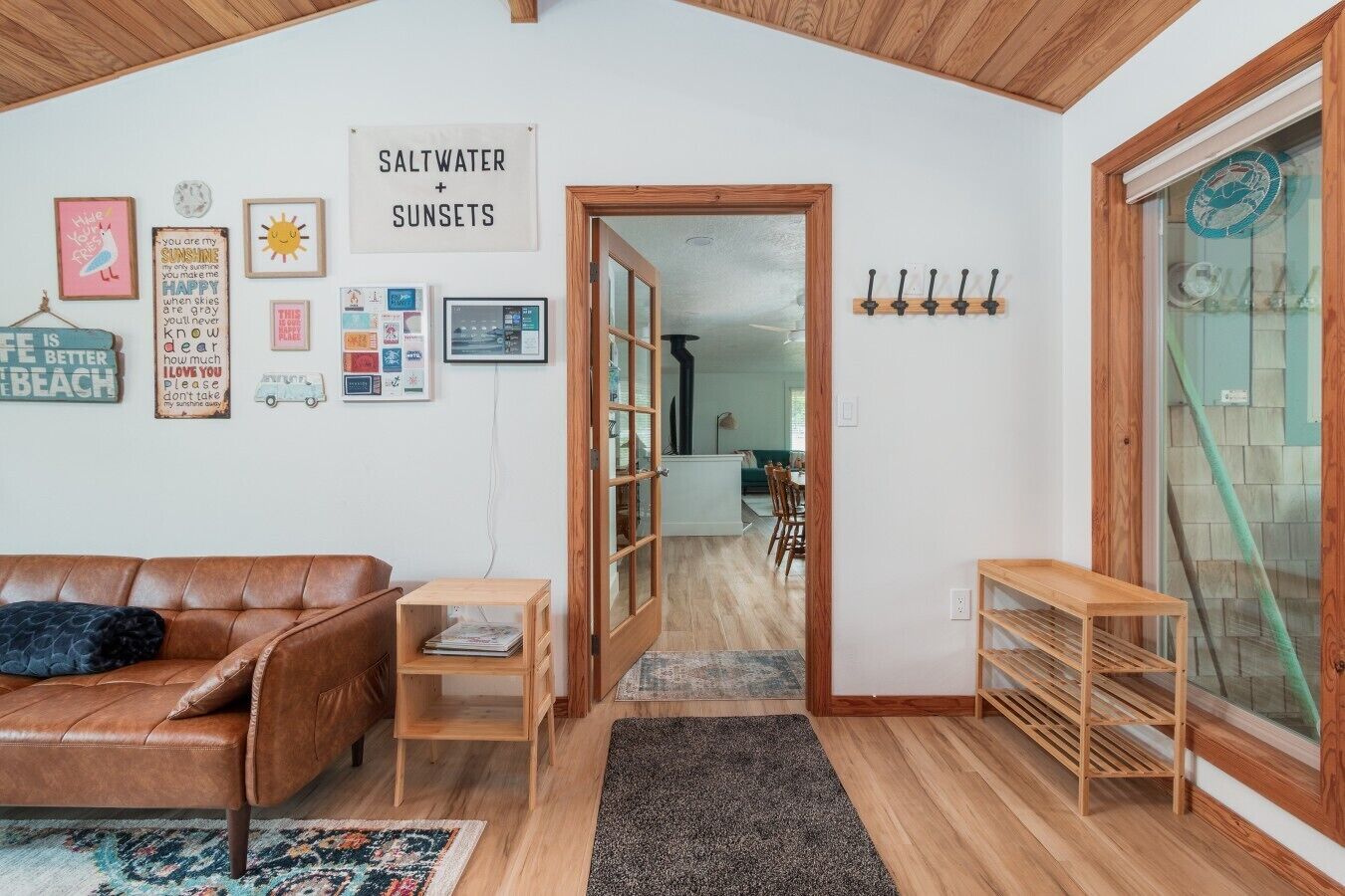 A cozy living room with a brown leather sofa, a wooden coffee table, and a bookshelf. A sign on the wall reads 'SALTWATER + SUNSETS.'