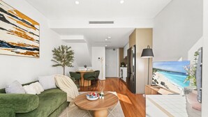Un saló modern amb un sofà verd, una zona de menjador amb taula i caires, una cuina amb nevera, i un quadre gran amb una escena de platja.