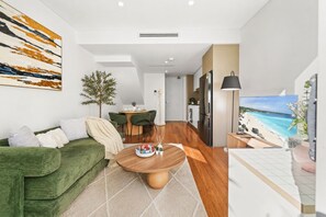 Un saló modern amb un sofà verd, una zona de menjador amb taula i caires, una cuina amb nevera, i un quadre gran amb una escena de platja.