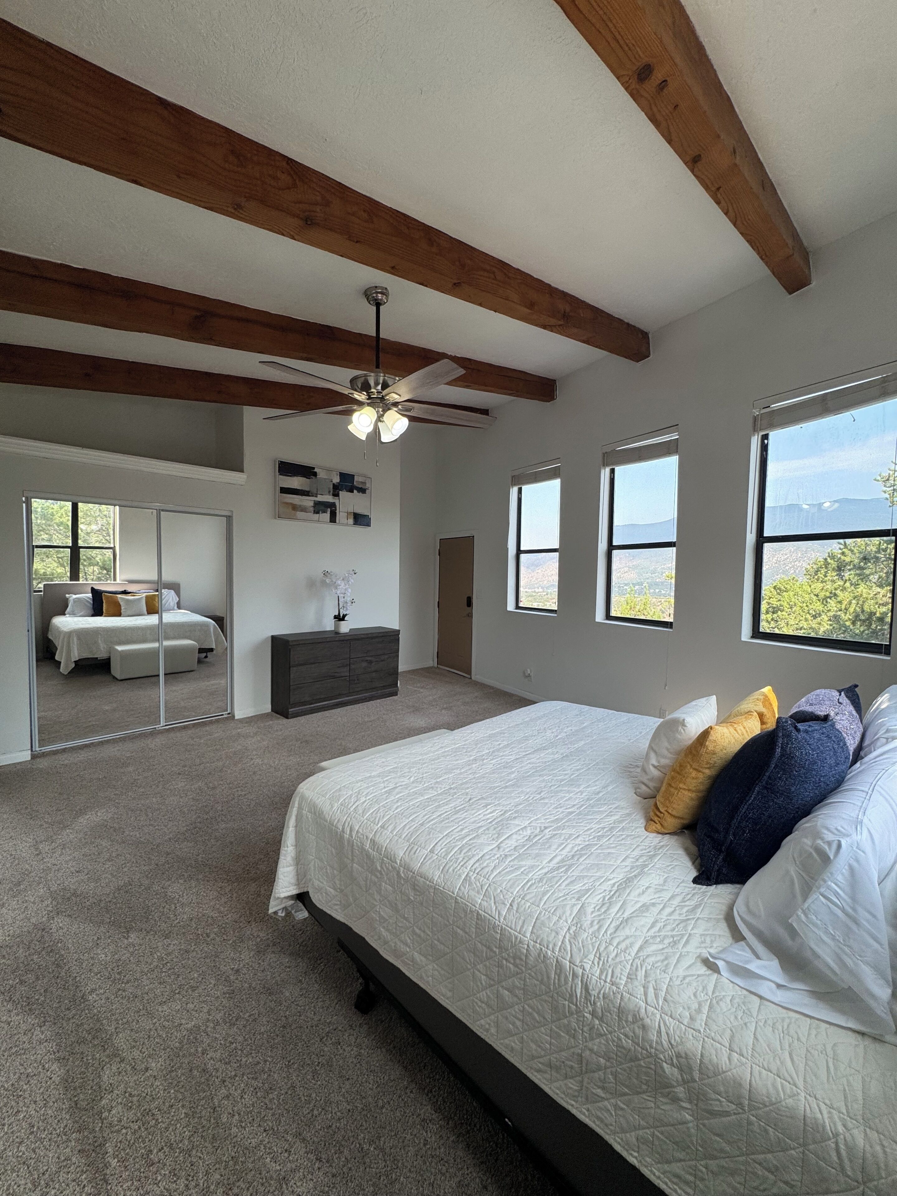 Une chambre avec un grand lit, un ventilateur de plafond, une commode et un miroir. Il y a des fenêtres donnant sur l’extérieur.