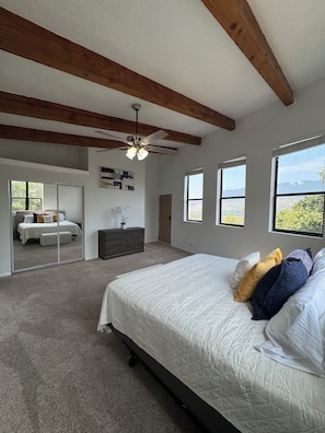 A bedroom with a large bed, a ceiling fan, a dresser, and a mirror. There are windows with a view of the outdoors.