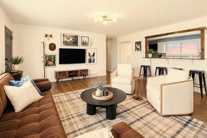 A living room with a brown leather sofa, a round coffee table, and a TV on the wall. There is a kitchen area with a window and stools. - Mins to RRG | Hot Tub, Game Room, Fire Pit (Stanton)