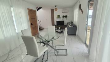 Een moderne eetruimte met een glazen tafel en stoelen, een bank en een televisie op de achtergrond.