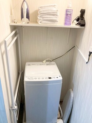 A compact laundry area with a washing machine, a stacked pile of white towels, a bottle of liquid soap, and an iron on a shelf. - Neo Universal 02 (Osaka, Konohana Ward, Nishikujo)