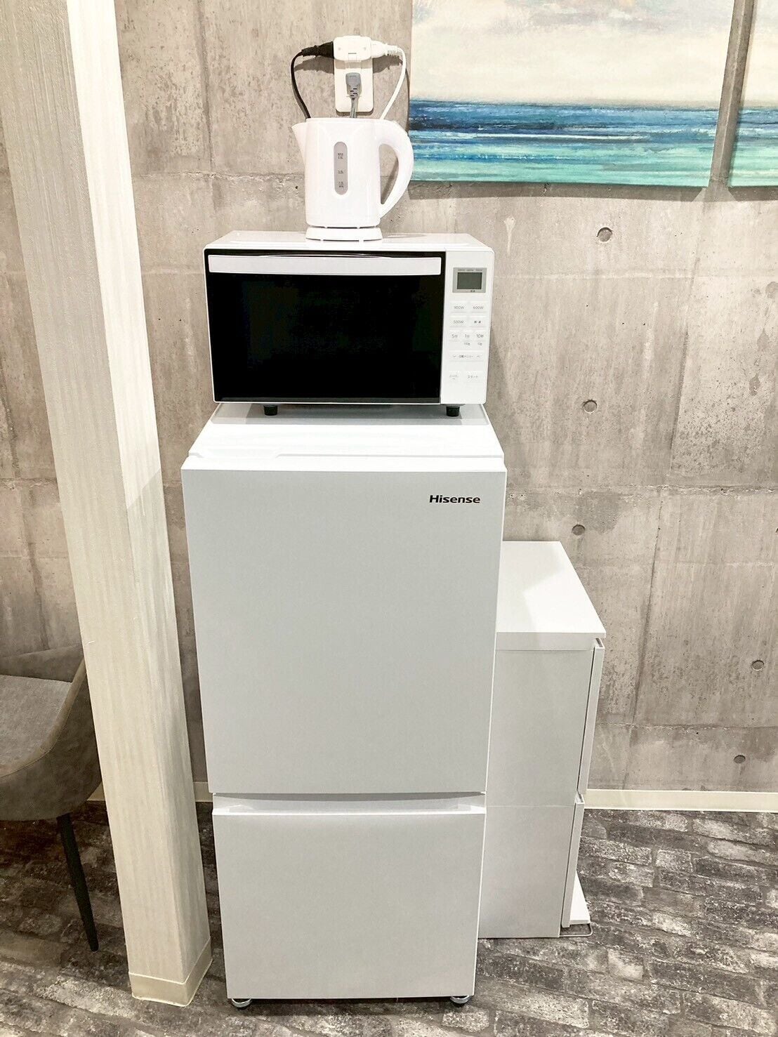 Un réfrigérateur Hisense surmonté d’un four à micro-ondes, placé contre un mur en béton orné d’un tableau représentant une scène de plage.