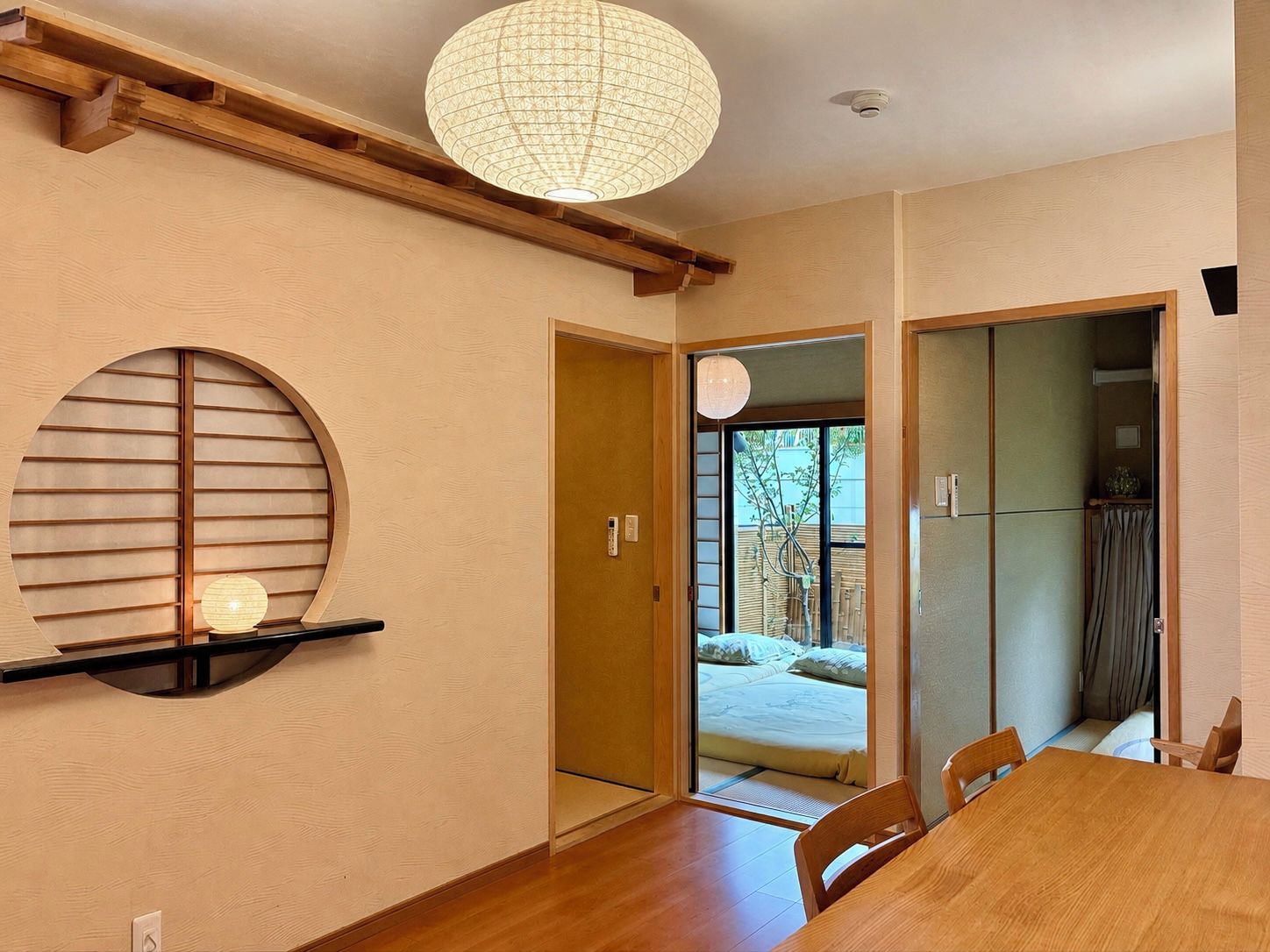 A room with a wooden dining table, chairs, a hanging light fixture, a sliding door leading to a bed, and a view of the outdoors.