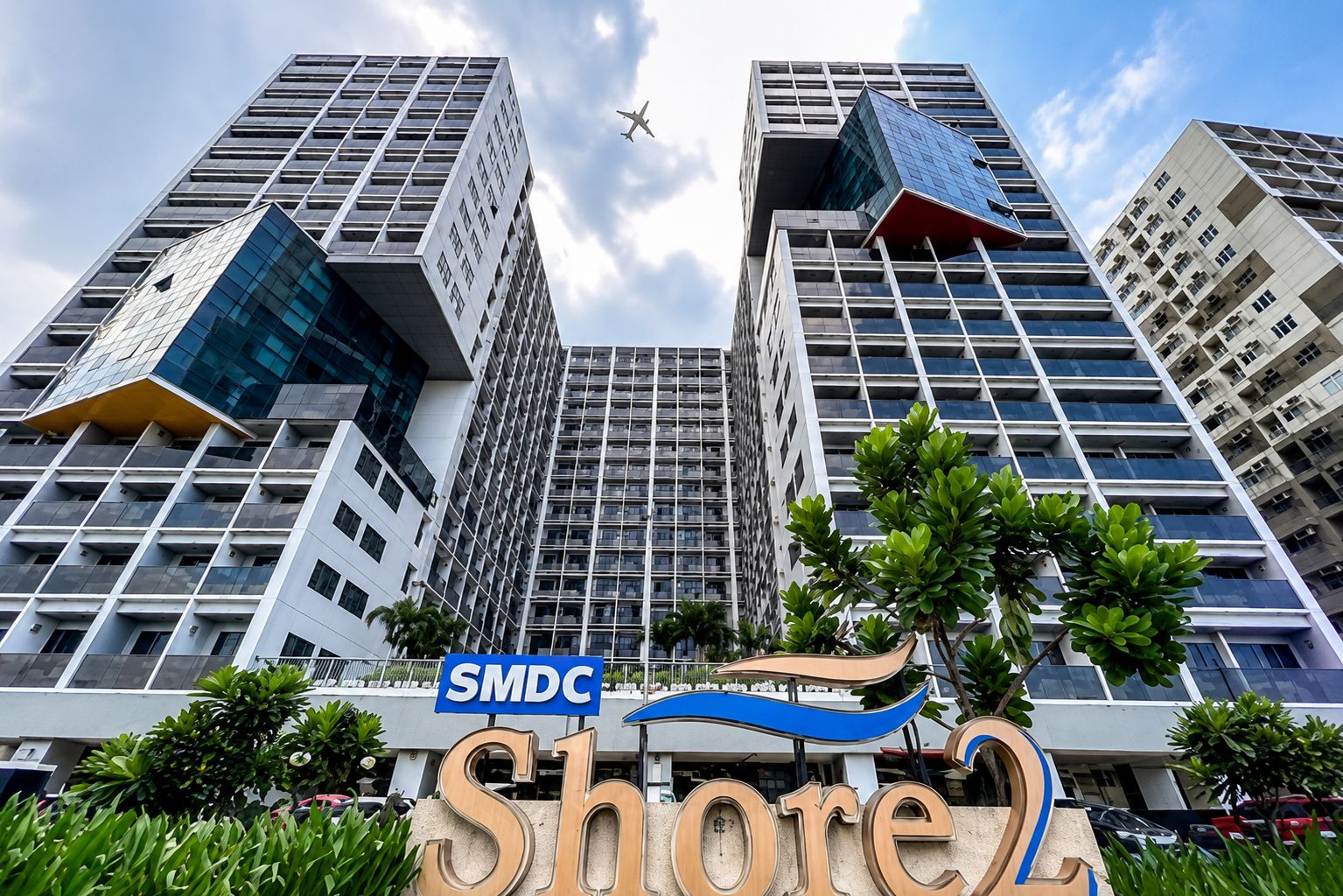 A modern building with a sign reading 'SMDC Share2' and a clear sky with a plane in the distance.