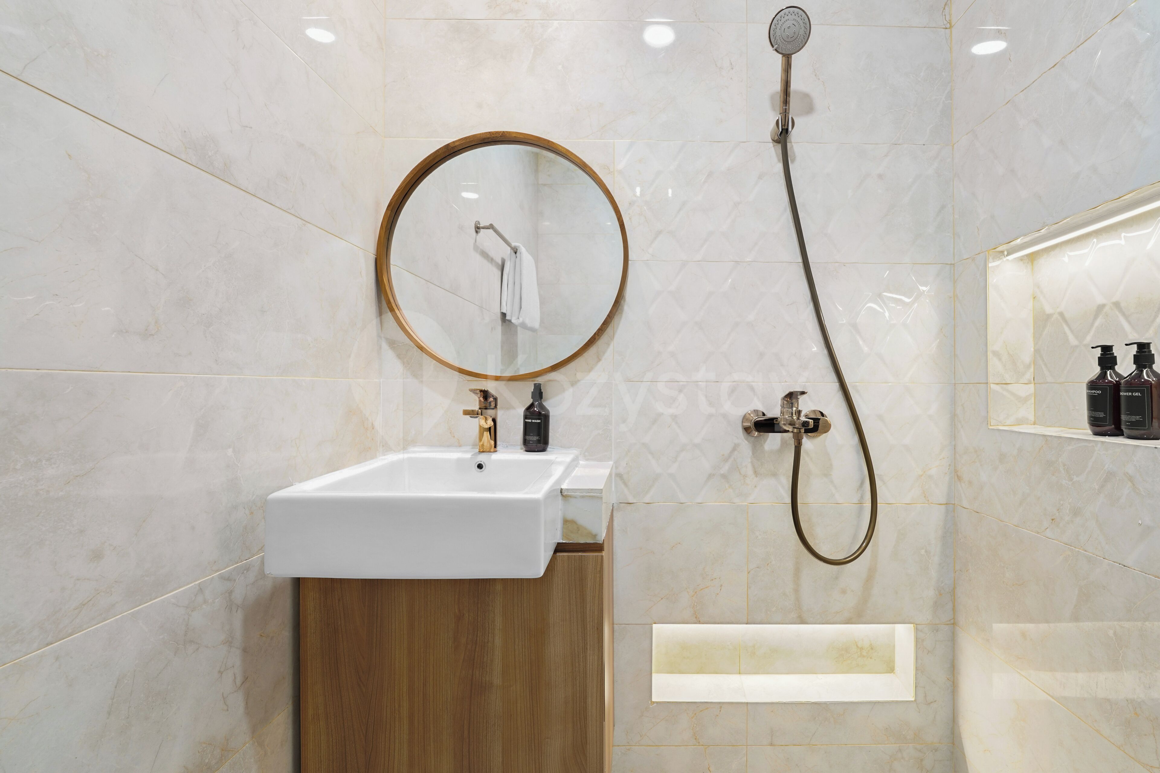 Un baño moderno con un lavamanos blanco, un espejo redondo, un cabezal de ducha manual y un gabinete de madera.