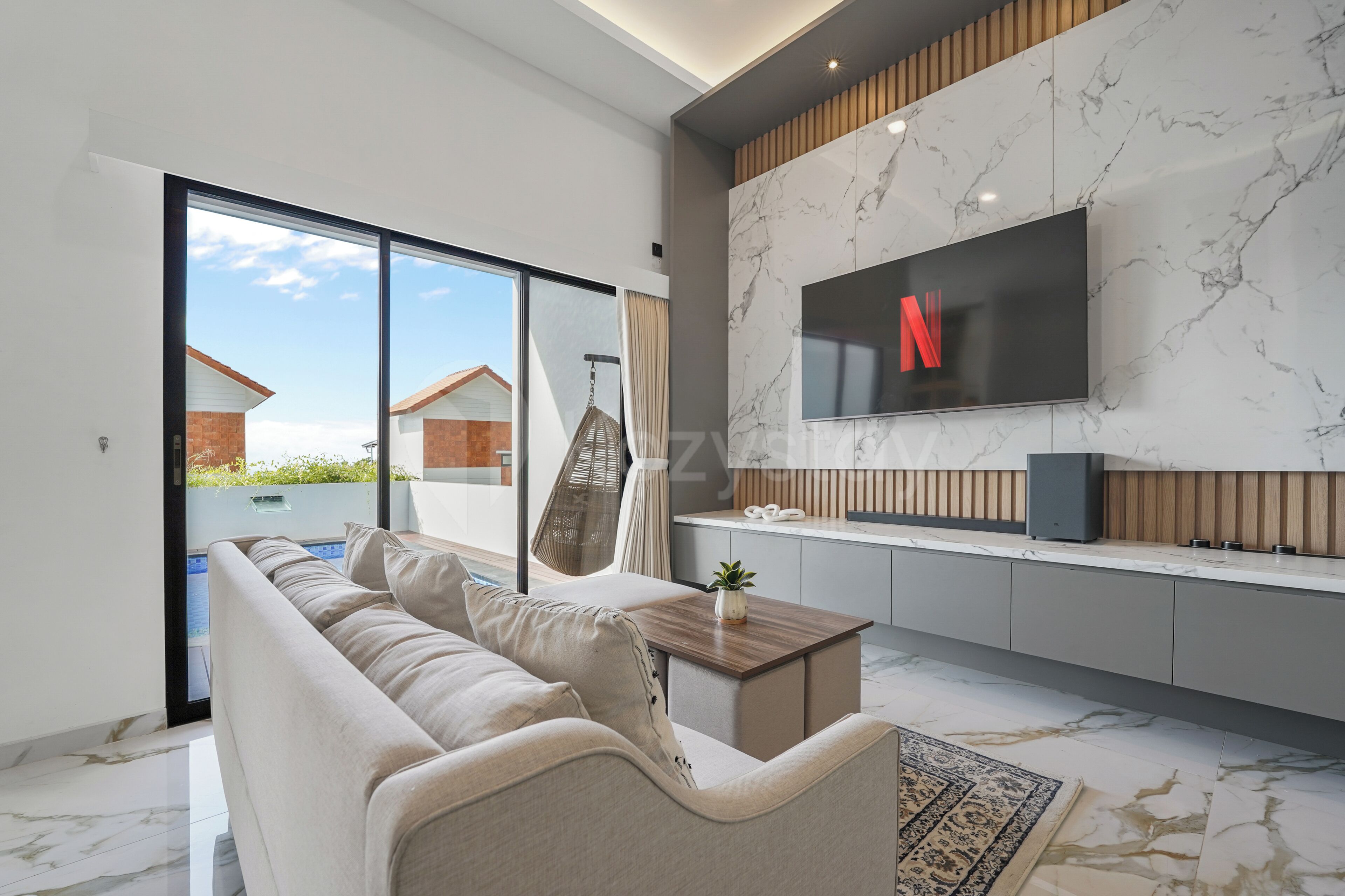 Ein modernes Wohnzimmer mit einer Couch, einem an einer Marmorwand montierten Fernseher und Blick auf einen Pool im Freien.