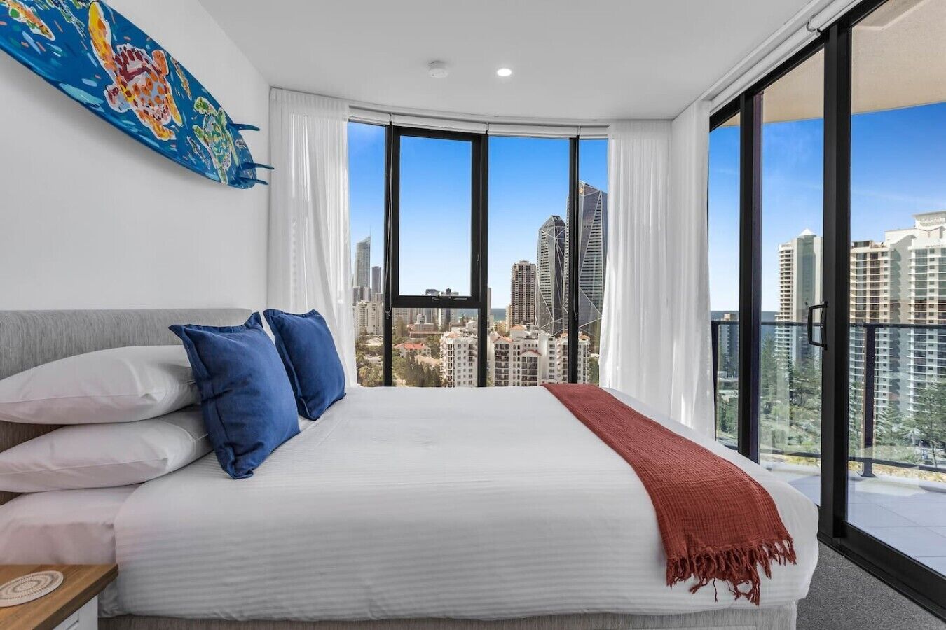 Camera d'albergo con un letto grande, biancheria da letto bianca e vista sulla città attraverso finestre a tutta altezza.