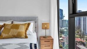 A bedroom with a bed, a nightstand, a lamp, and a large window with a city view. - Lvl 15 Luxury 2 Bedroom APT with Beach Views (Broadbeach)