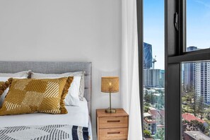 Ein Schlafzimmer mit einem Bett, einem Nachttisch, einer Lampe und einem großen Fenster mit Blick auf die Stadt.