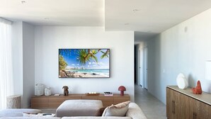 A modern living room with a large flat-screen TV mounted on the wall, a wooden cabinet, and a sofa with cushions. - Lvl 15 Luxury 2 Bedroom APT with Beach Views (Broadbeach)