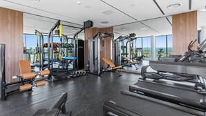A modern gym with various exercise equipment, large windows, and a city view. - Lvl 15 Luxury 2 Bedroom APT with Beach Views (Broadbeach)