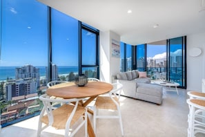 A modern living room with a city view, a sofa, a dining table, and a large window. - Luxury L16  2 bedroom APT with Beach Views (Broadbeach)