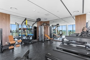 A modern gym with various exercise equipment, large windows, and a city view. - L18 Stunning 3 BRM Apt with Beach Views (Broadbeach)